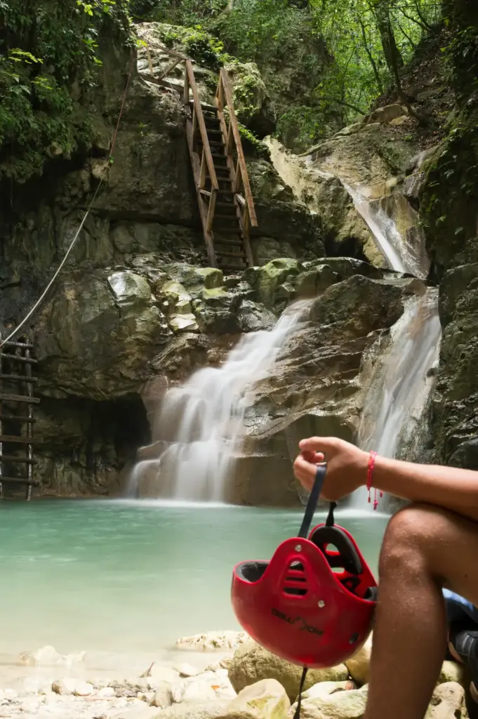 Visitante con casco rojo en la mano frente a una cascada y poza turquesa en Damajagua